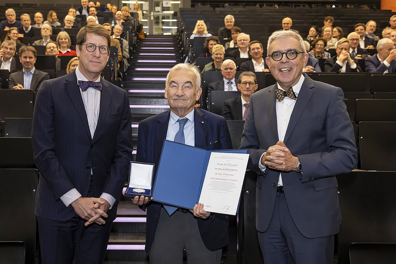 Auf dem Bild stehen drei Personen. Prof. Ertl in der Mitte, der eine Urkunde und eine Medaille in seinen Händen hält. Im Hintergrund gehen die Treppen hoch zum Hörsaal. Rechts und links sitzen viele Leute.