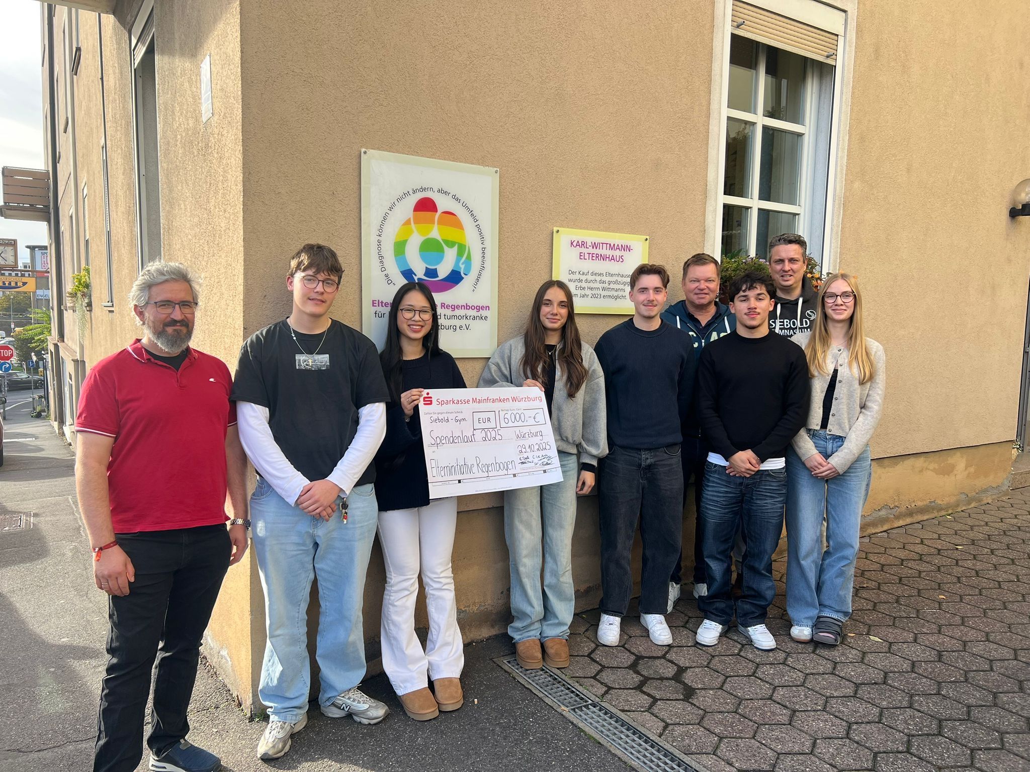 Auf Bild stehen mehrere Schüler mit Lehrern vor dem Gebäude der Elterninitiative. In der Mitte halten zwei Schüler einen großen Scheck in der Hand. Im Hintergrund sind am Haus zwei Schilder zu sehen. Elterninitiative Regenbogen und Karl-Wittmann-Elternhaus..as eine Schi