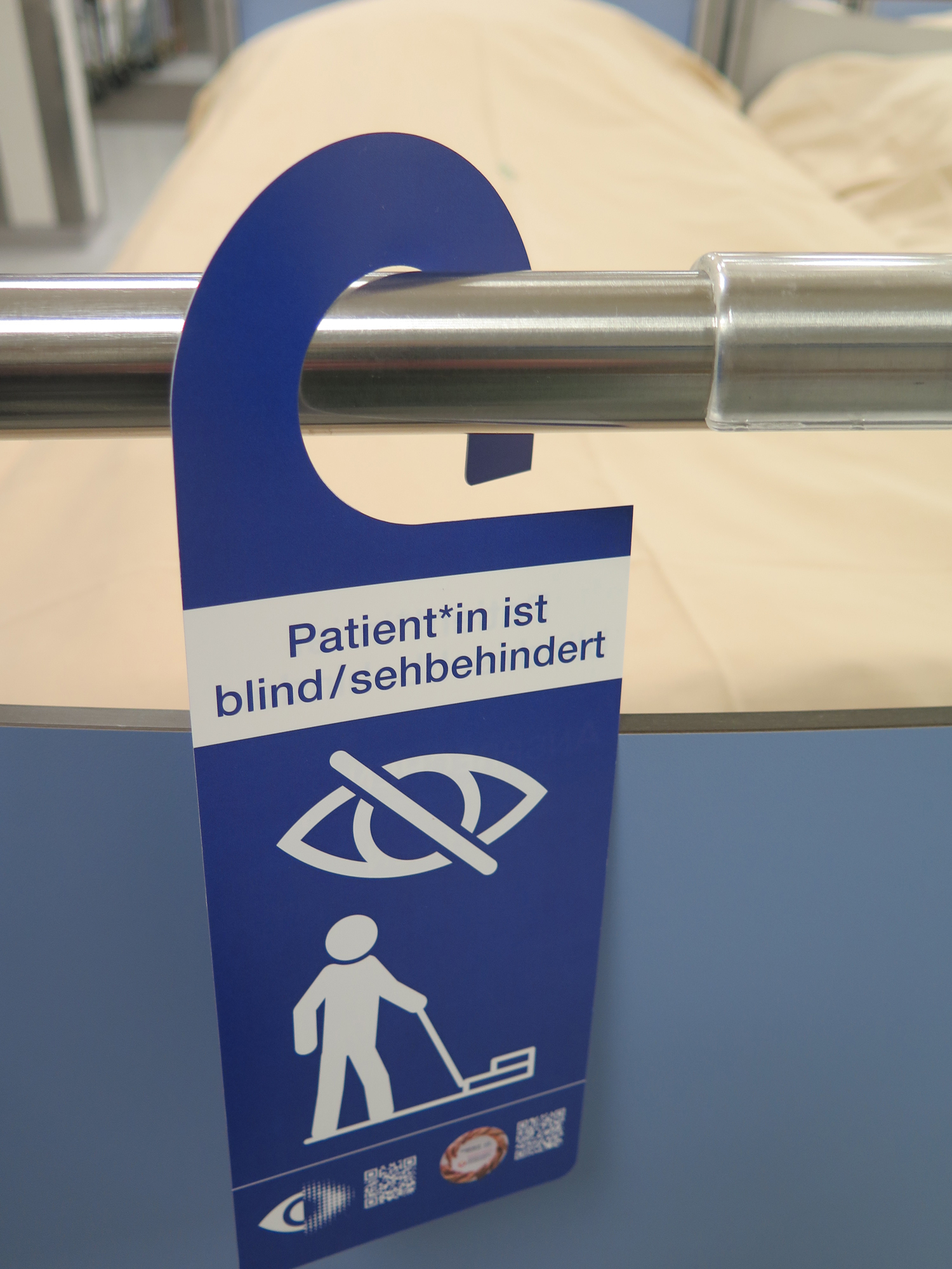 Auf dem Bild sieht man ein frisch bezogenes Krankenbett an dem am Ende des Fußes ein blauer Anhänger am Gestänge hängt. Auf dem steht Patient*in ist blind / sehbehindert. Darunter sind zwei Piktogramme. Ein durchgestrichenes Auge und ein Strichmännchen mit Stock, der vor einer Stufe steht. 