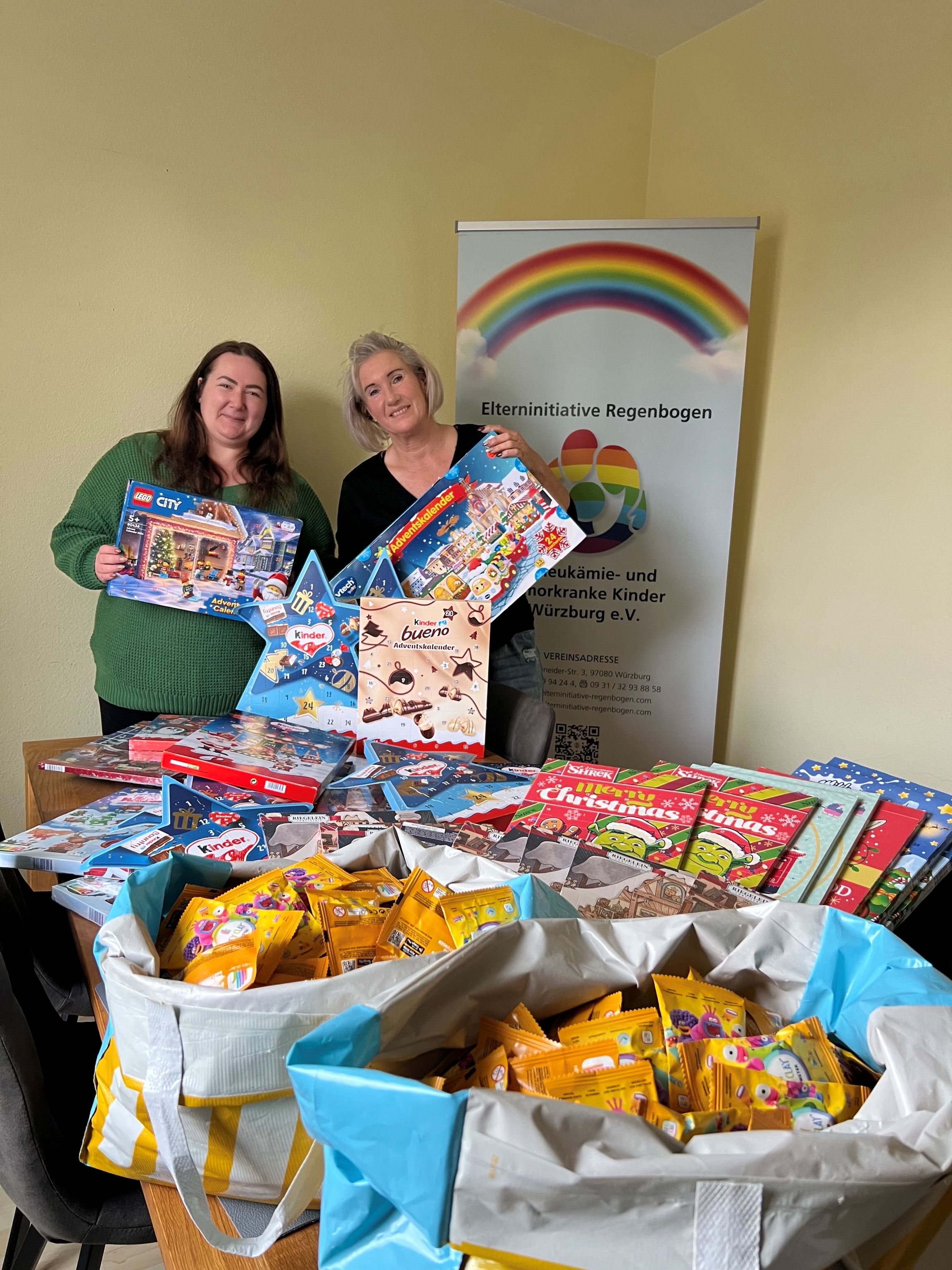 Auf dem Bild sind zwei Frauen zu sehen, die in ihren Händen verschiedene Adventskalender halten. Vor ihnen auf dem Tisch stehen mehrere Tüten mit verschiedenen Sachspenden. Im Hintergrund steht ein Roll-up der Elterninitiative Regenbogen.