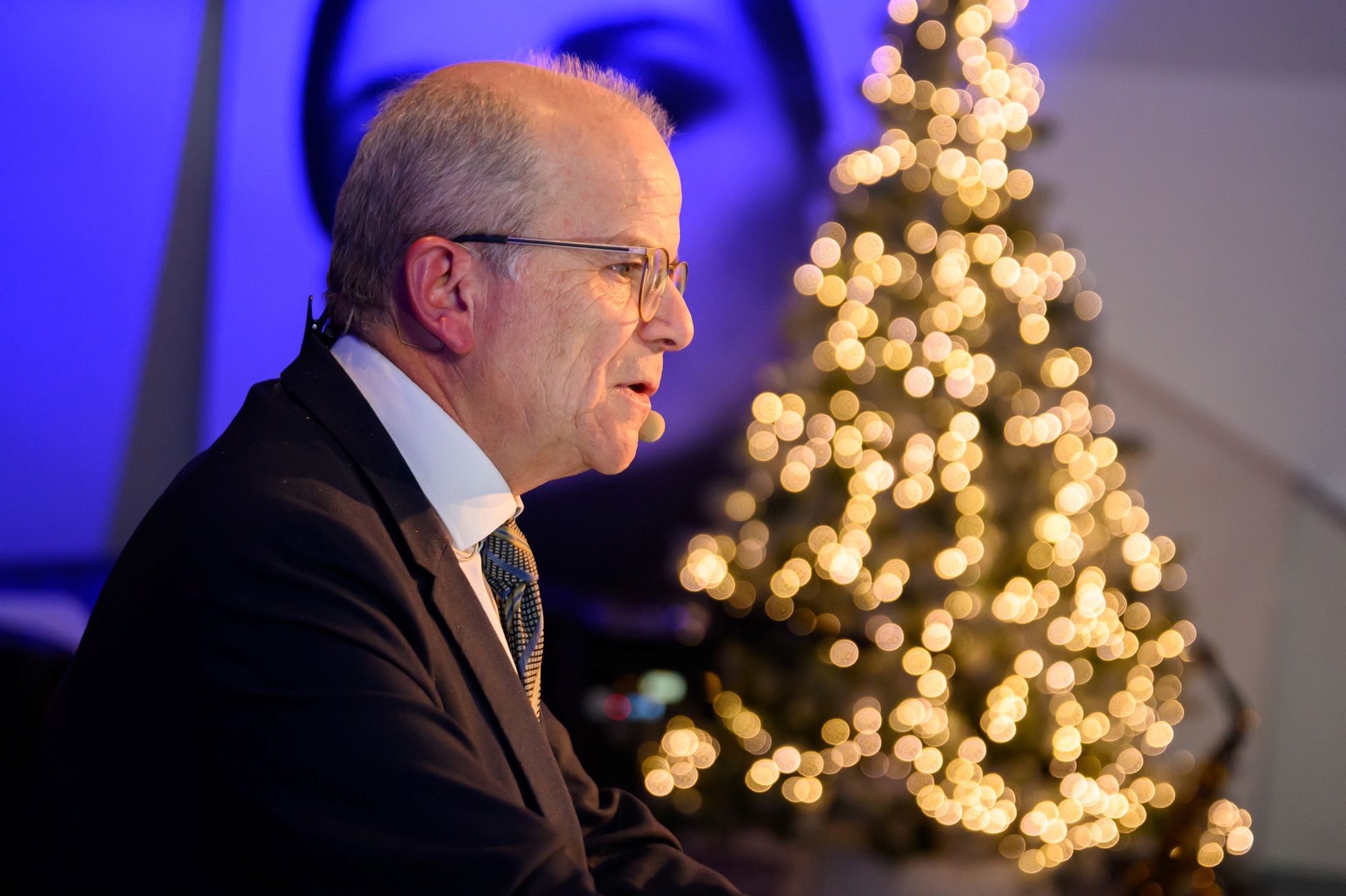Auf dem Bild sieht man einen Mann von der Seite, der eine Rede hält. Im Hintergrund leuchtet ein Weihnachtsbaum. 