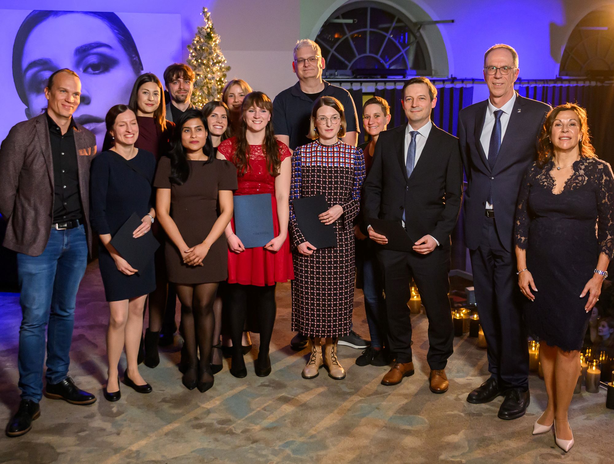 Auf dem Bild sieht man Gruppenbild des Forschungsteam. Ein Teil hält eine schwarze Mappe in den Händen.  Der Hintergrund leuchtet blau. Auf der linken Seite ist ein großes Poster mit einem Frauenkopf zu sehen. Ebenso ein leuchtender Weihnachtsbaum sowie ein Bogenfenster.