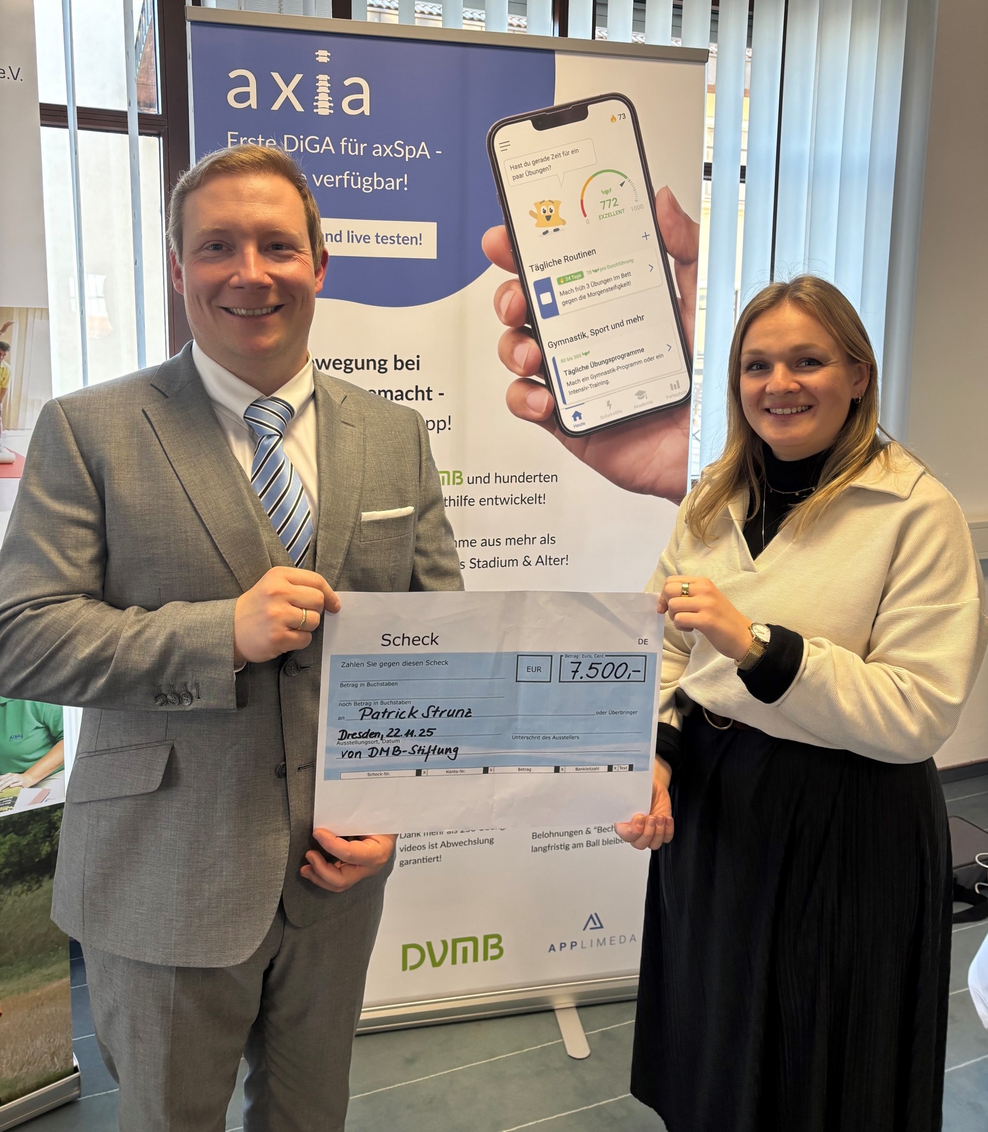Der Gewinner und die Spenderin halten einen auf DinA2 ausgedruckten Scheck hoch, dahinter ein Rollup von der App Axia.  