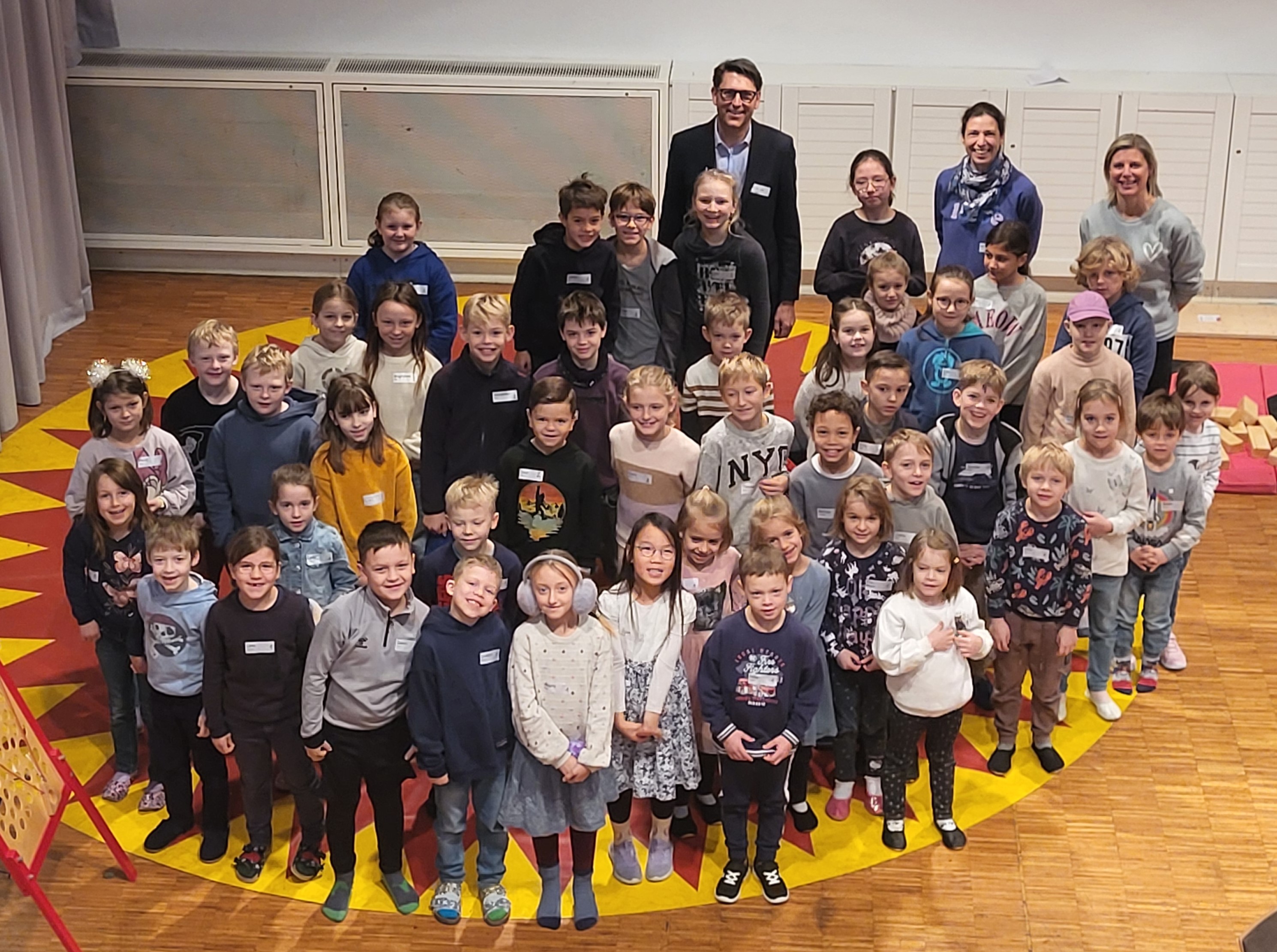 Das Bild wurde von oben runter auf eine Schar von Kindern fotografiert, die auf einem runden gelben Teppich mit roten Zacken stehen. Im Hintergrund der Kinder stehen der Ärztliche Direktor und zwei Resilienztrainerinnen. 