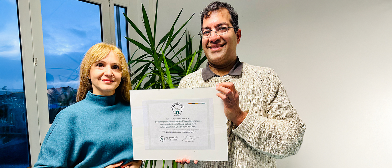 Bild zeigt eine Frau und einen Mann, die vor einem Fenster und einer Palme stehen und in der Hand eine Nachhaltigkeits-Zertifizierung halten. 