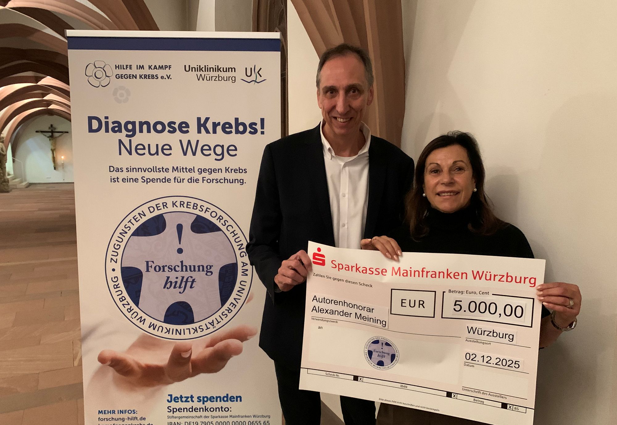 Foto zeigt Spendenübergabe. Prof. Meining und Frau Nelkenstock halten einen Scheck über 5.000 Euro in der Hand. Links im Hintergrund steht ein Roll-up mit Diagnose Krebs - Neue Wege mit dem Logo Forschung hilft.