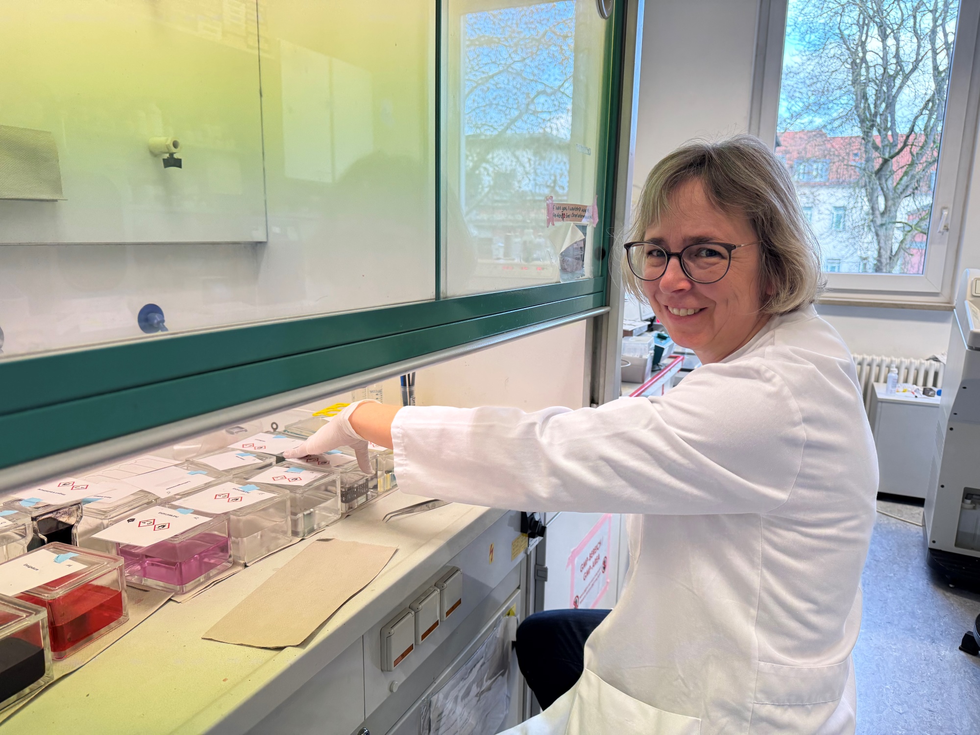 Die Technische Angestellte sitzt im Laborkittel vor der Laborbank vor Gefäßen mit unterschiedlichen Färbungenfäße