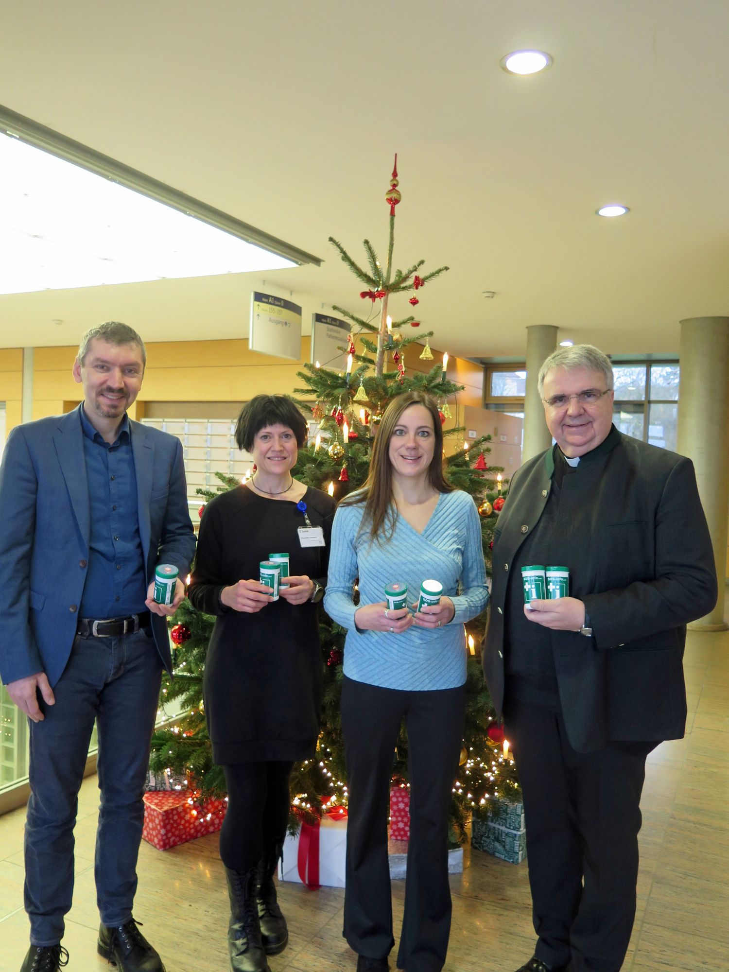 Die Notfalldosen werden von vier Personen präsentiert. Im Hintergrund steht ein Weihnachtsbaum. 