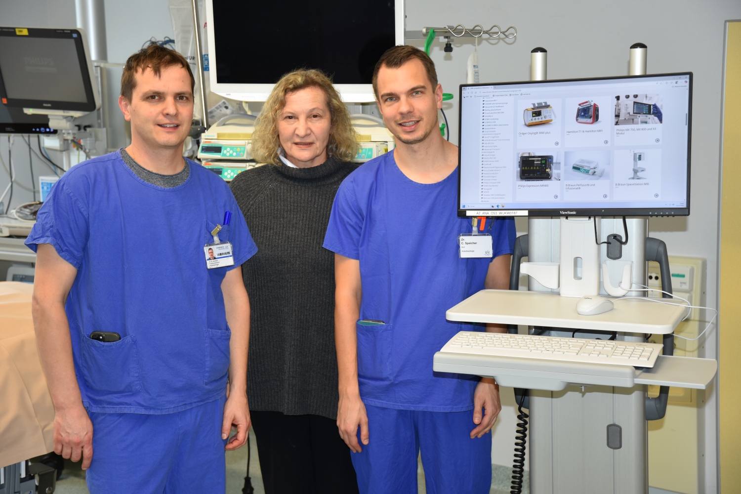Die beiden Ärzte stehen in blauer Arbeitskluft rechts und links von Anna Kellersmann, daneben ein medizinisches Gerät. 