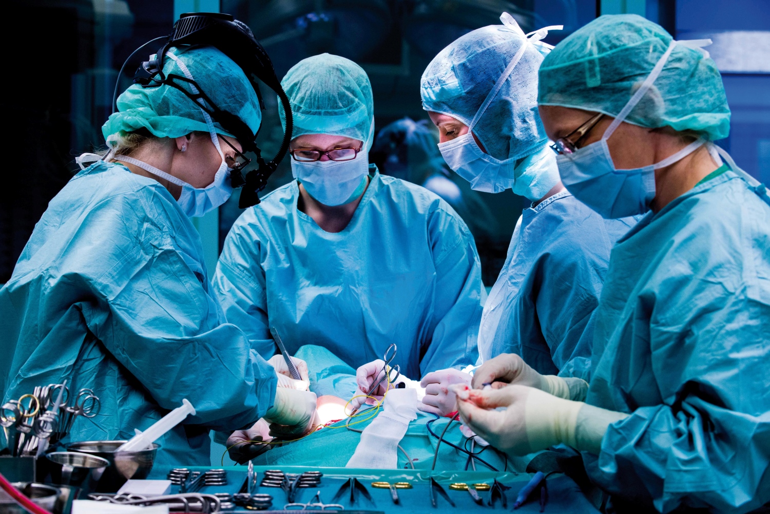 4 Fachkräfte in OP-Kluft mit Kittel, Haube und Mundschutz stehen am Operationstisch und behandeln einen Patienten
