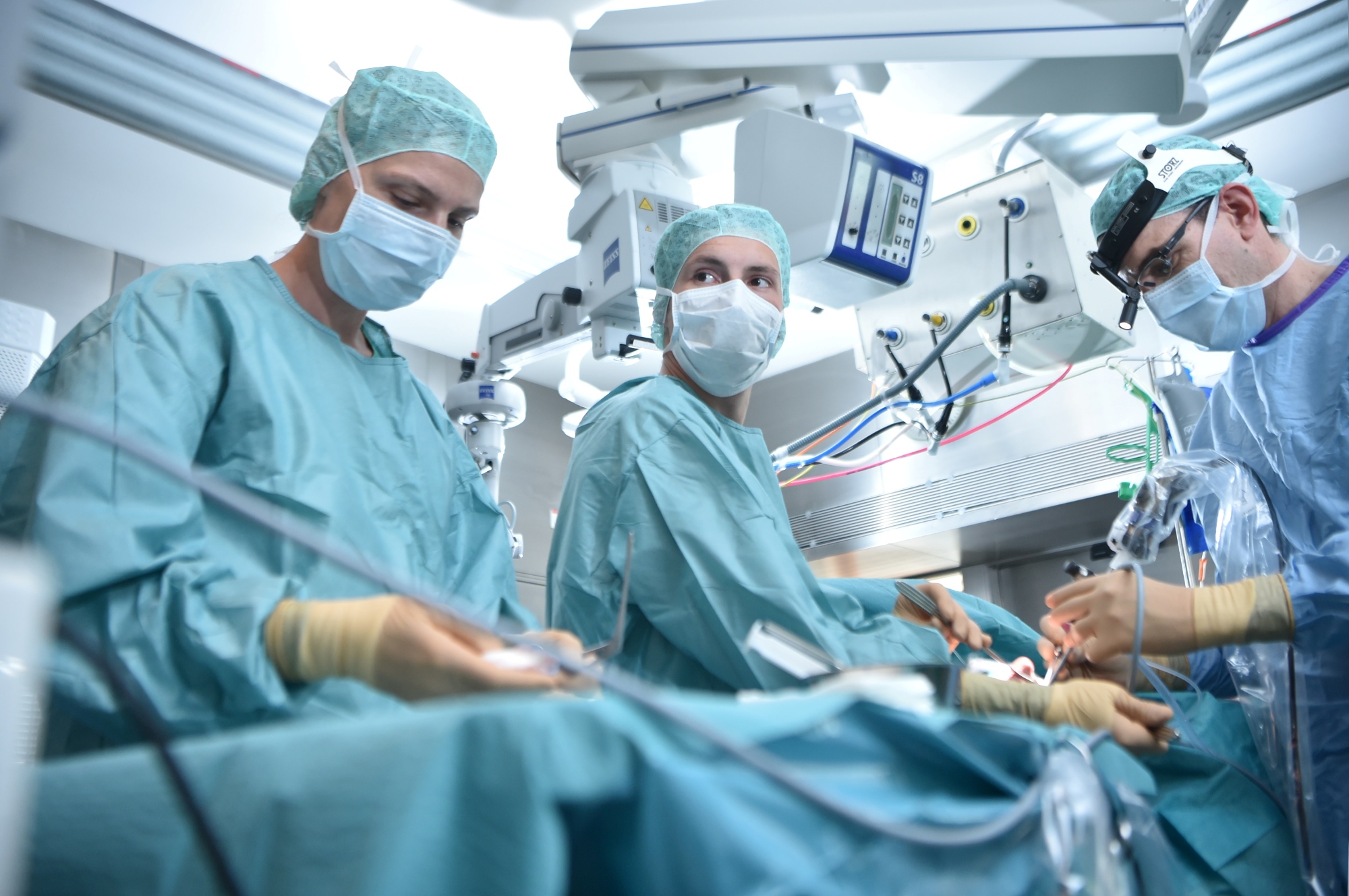 Drei Fachkräfte in OP-Kluft mit Kittel, Haube und Mundschutz stehen am Operationstisch und behandeln einen Patienten