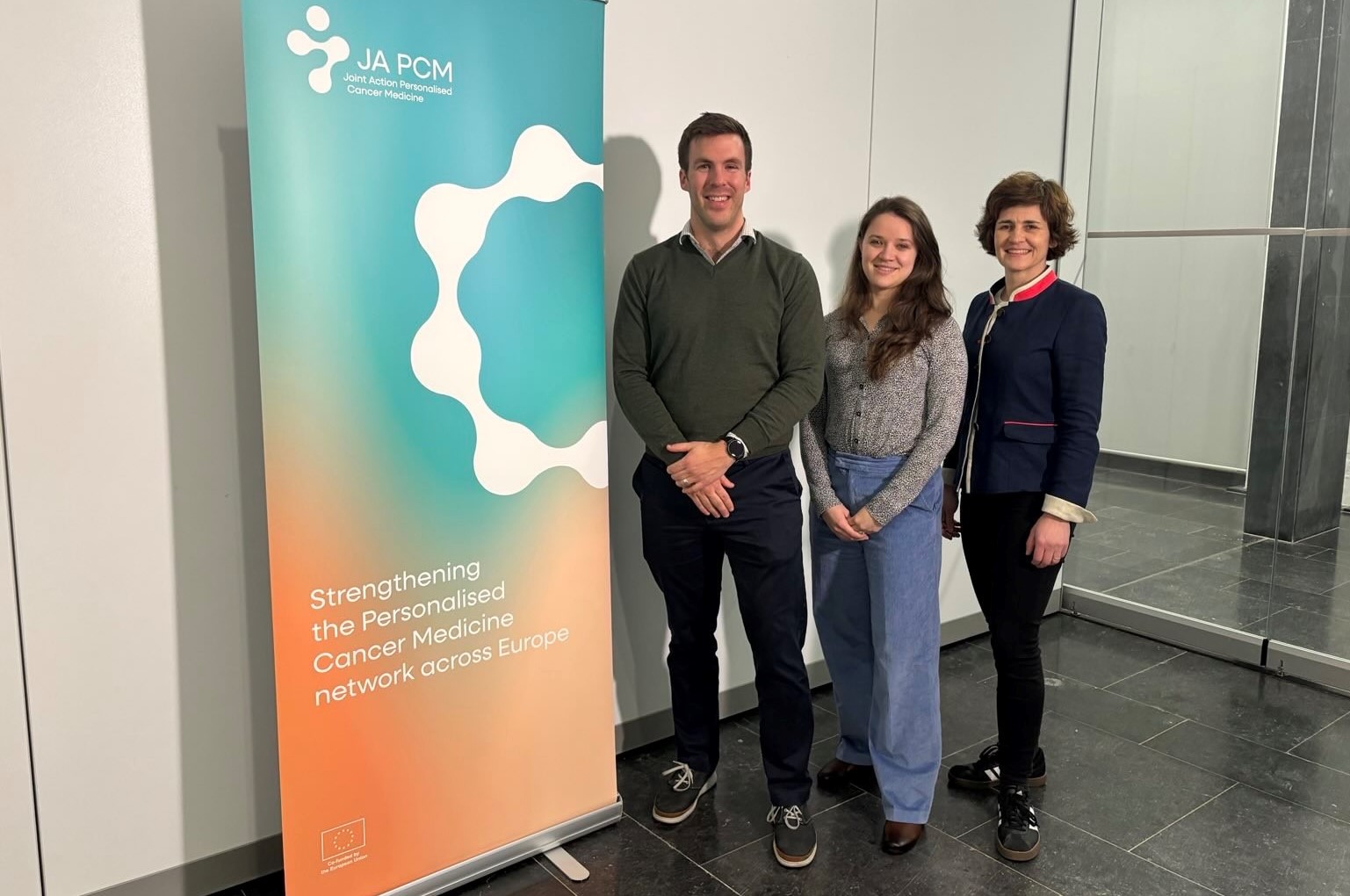 Die drei Forschenden stehen neben einem Roll-up der Joint Action, auf dem der Slogan steht "Strengthening the Personalised Cancer Mecicine Network across Europe 