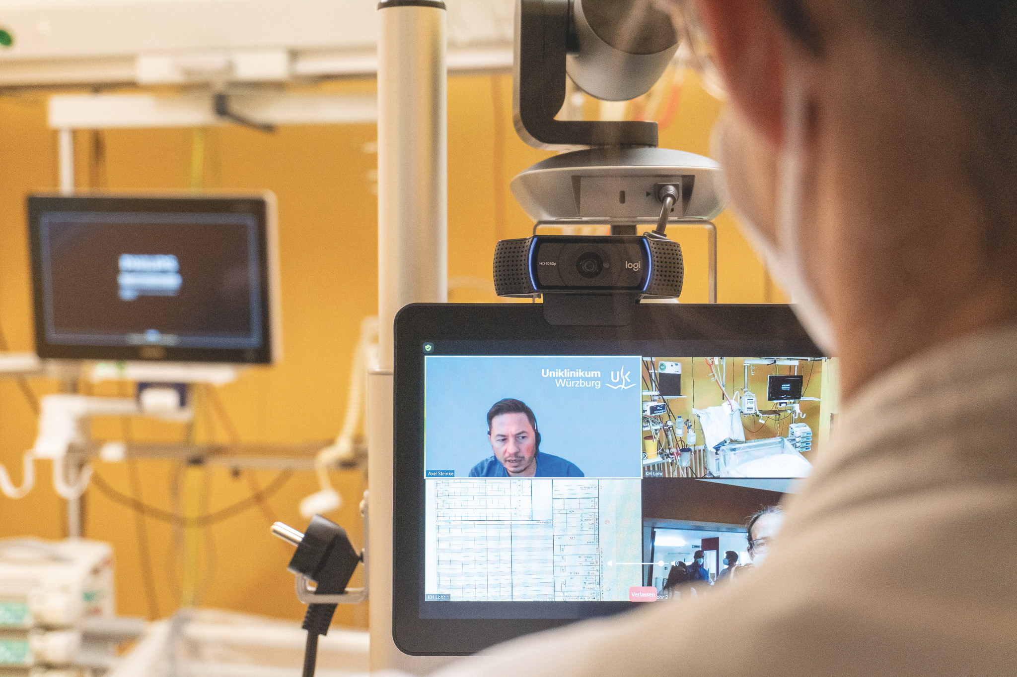 Hintere Kopfansicht einer Person, die auf einen Monitor schaut ,auf dem ein Arzt vom UKW zu ihr spricht. In weiteren Feldern auf dem Monitor sind das Patientenbett und Parameter zu sehen.   