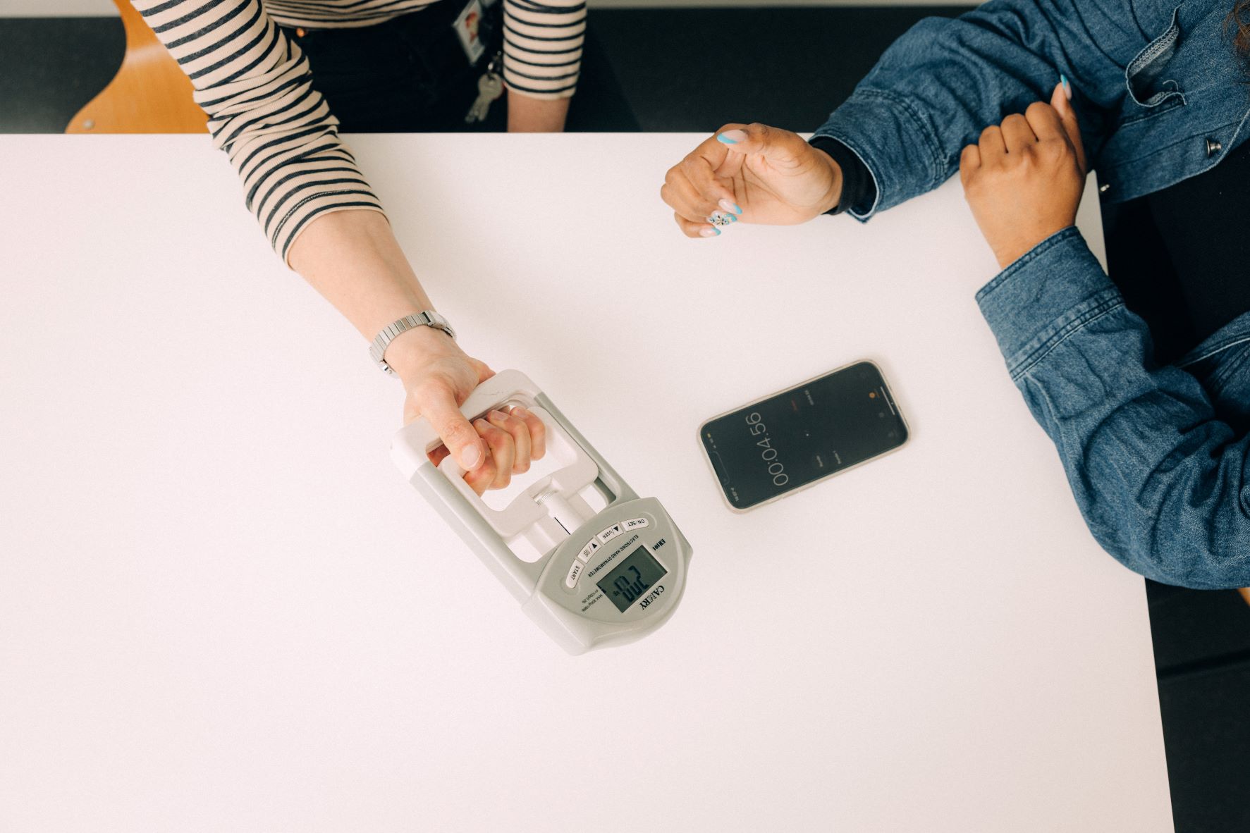 Szene einer Messung am Tisch: eine Person hält den Manometer, eine andere sitzt daneben und hat Smartphone vor sich liegen, vermutlich wird die Zeit gestoppt. 