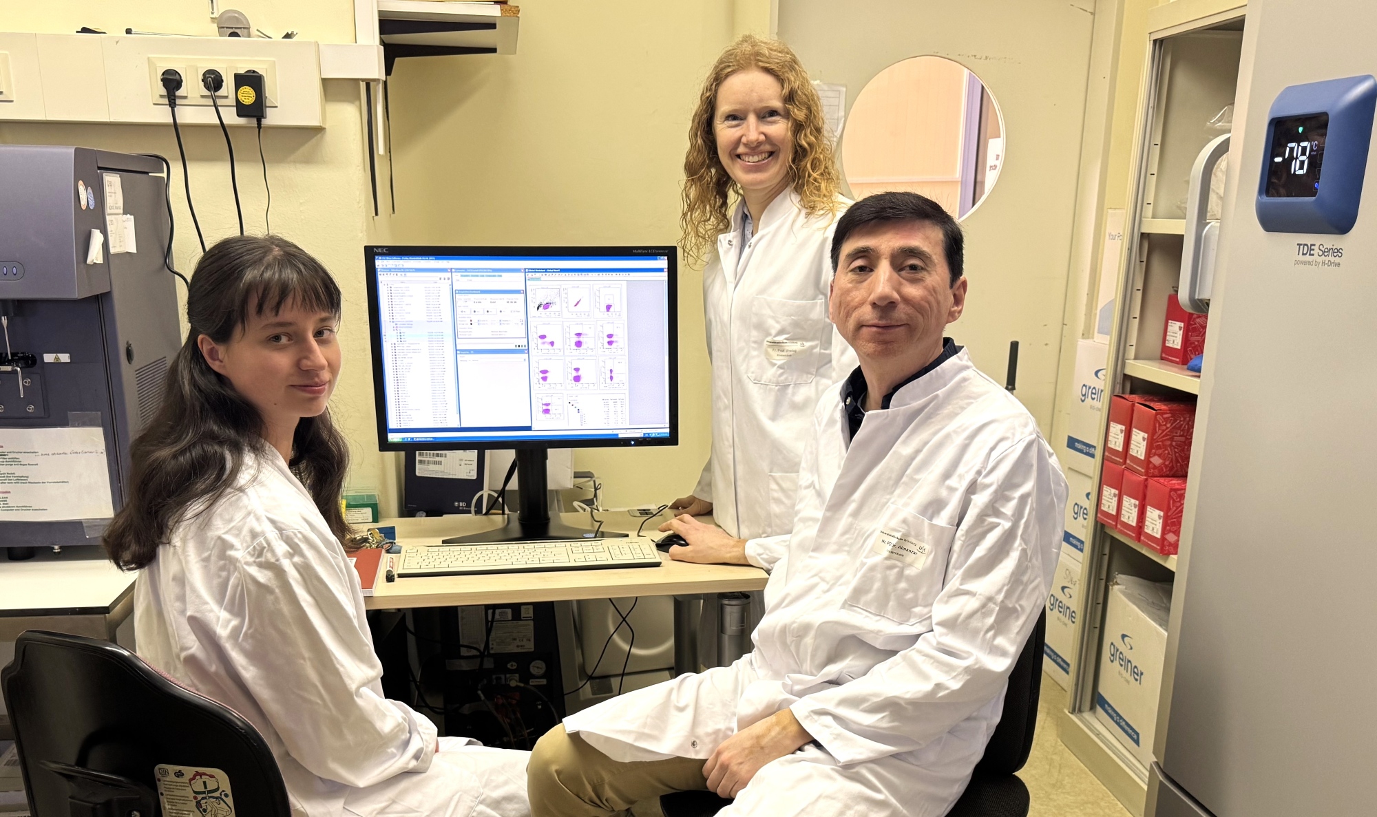 Die drei Forschenden sind im Labor, zwei sitzen, Martina Prelog steht, alle blicken in die Kamera, im Hintergrund ist ein Monitor, der die aktuelle Studie zeigt. 