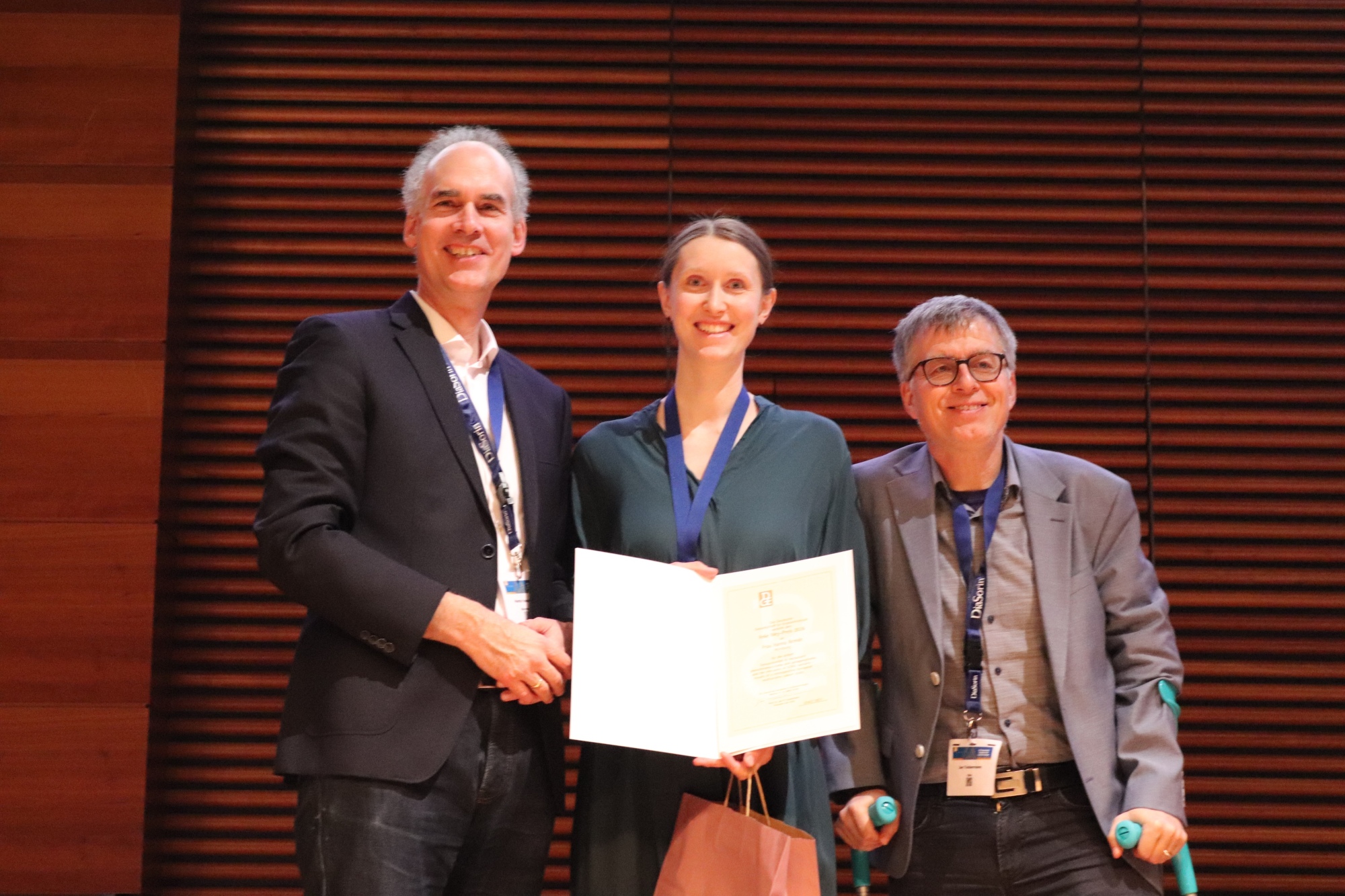 Preisträgerin mit Urkunde auf der Bühne, links steht Martin Fassnacht, rechts von ihr Jan P. Tuckermann mit Krücken. 