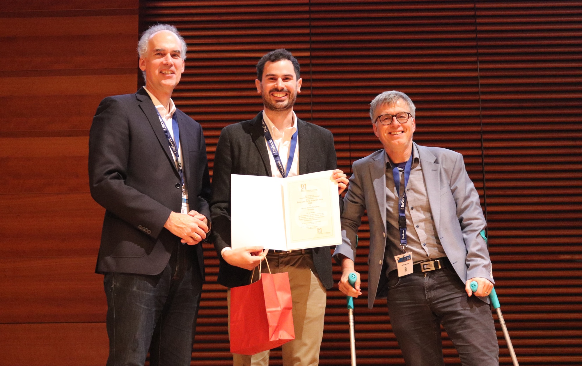 Preisträger Dr. Mario Detomas mit Urkunde auf der Bühne, rechts steht  Prof. Jan P. Tuckermann auf Krücken, links von ihm Prof. Martin Fassnacht.
