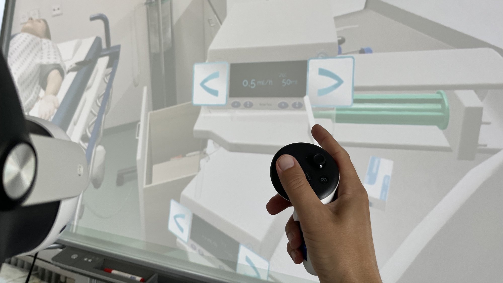 VR-Situation aus Sicht des Anwenders: links im Bildrand die VR-Brille und in der Mitte des Bildes die Hand des Anwenders mit VR-Gerät - im Hintergrund die virtuelle Welt mit Patient und Geräten