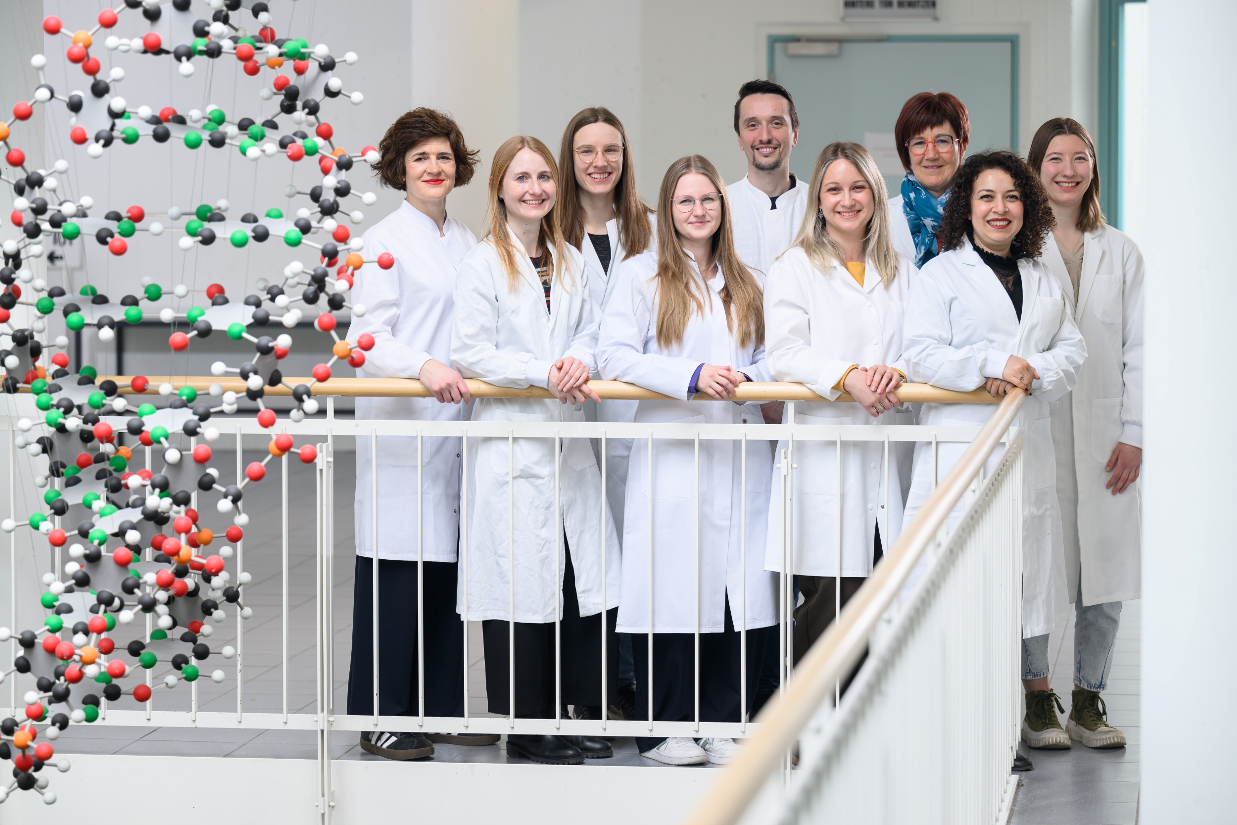 Die neun Mitarbeitende des Referenzdiagnostik-Labors stehen in weißen Kitteln im Foyer des Biozentrums, daneben hängt ein DNA-Mobile. 