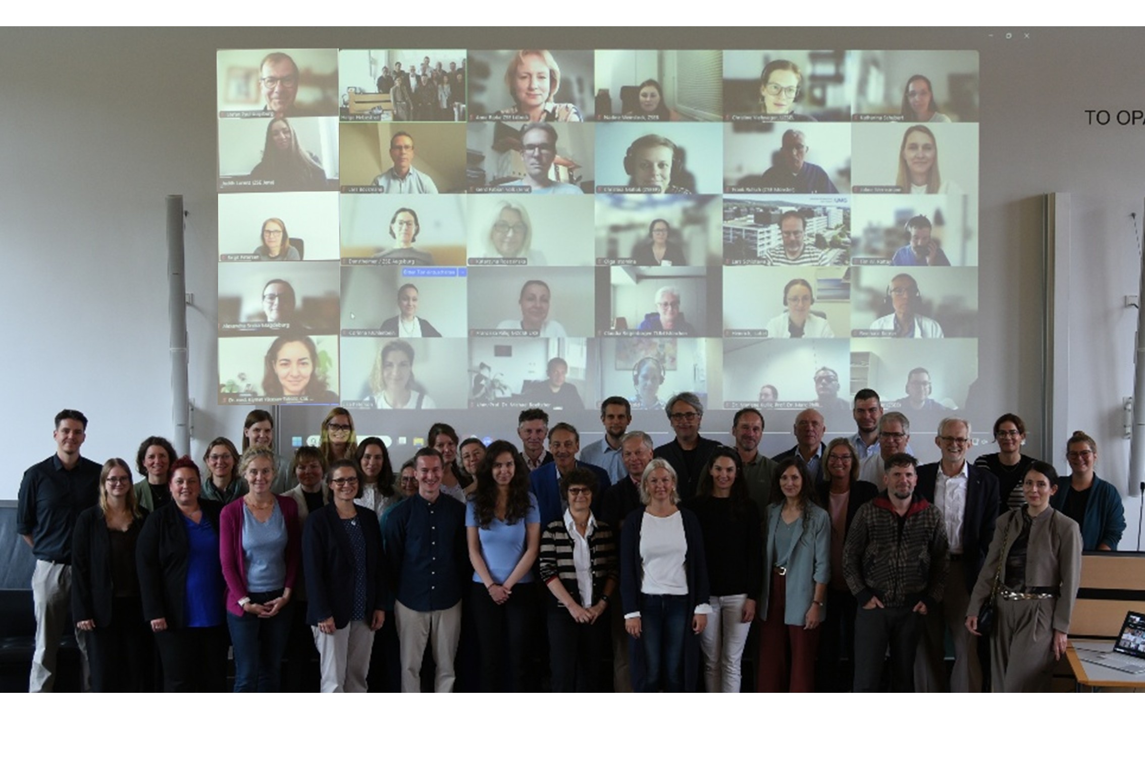 Gruppenfoto nach Verabschiedung der Satzung. Im Hintergrund sind an einer Wand die virtuellen Teilnehmer zu sehen. 