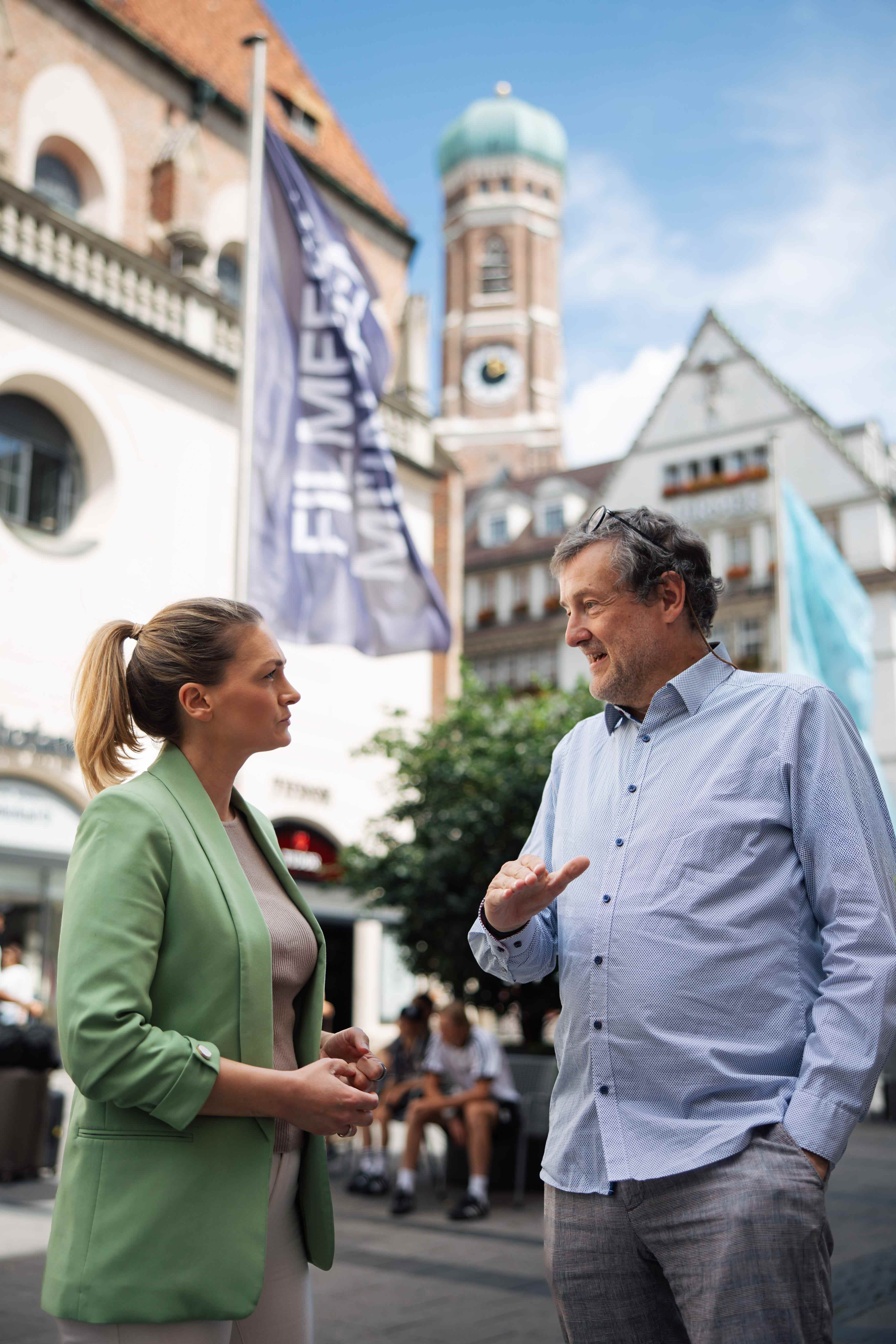 Bayern Gesundheitsministerin Judith Gerlach bei einem Gespräch mit einem Mann. Im Hintergrund sieht man eine Fahne und Münchens Kirche.