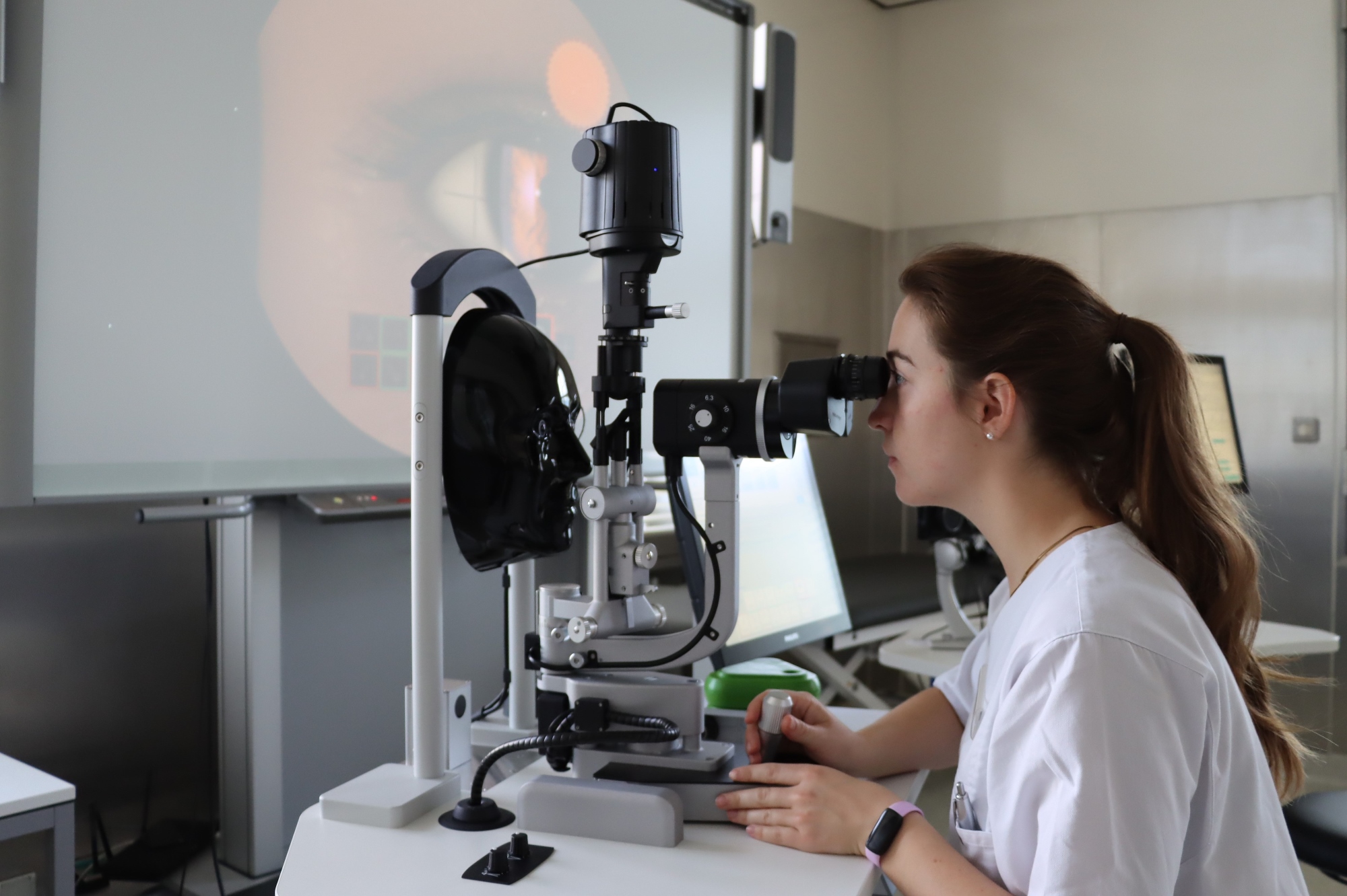 Studierende sitzt am Simulator für die Spaltlampe - der Patient ist ein schwarzes Kopfmodel. Im Hintergrund ist das Auge in groß auf einem Monitor zu sehen. 