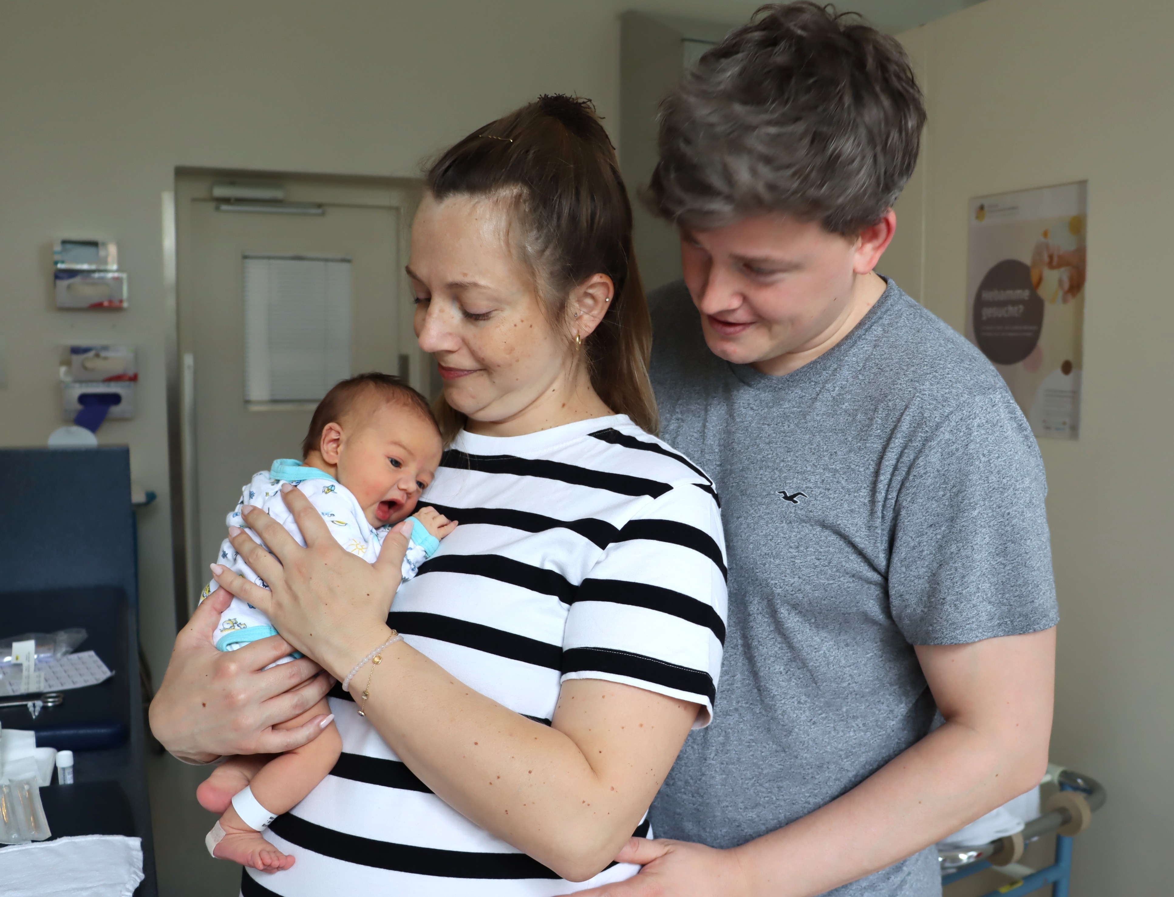 Die Mutter hält den Säugling auf dem Arm, der Vater steht daneben. 