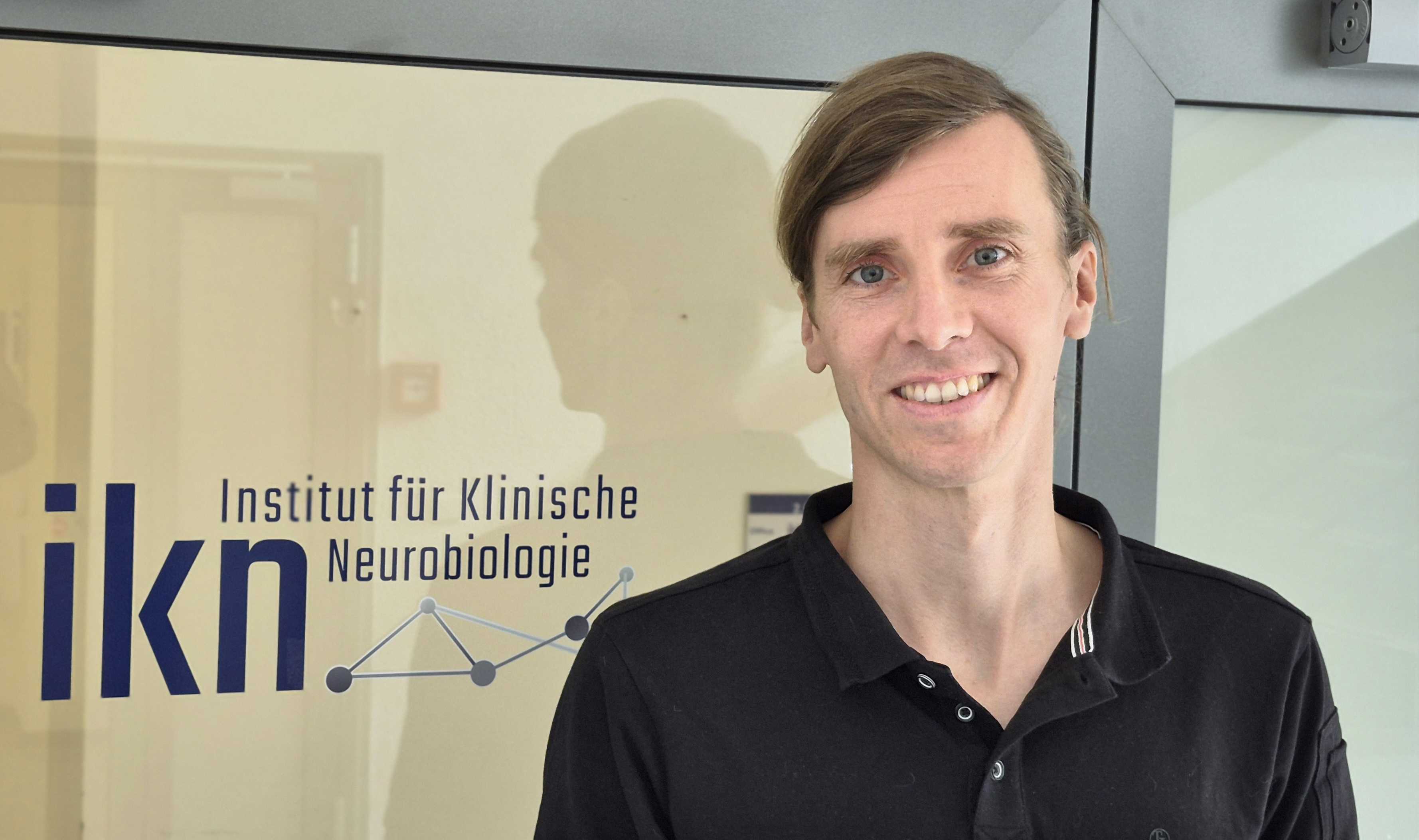 Dominik Groos vor einer Glastür mit der Beschriftung ikn Institut für Klinische Neurobiologie