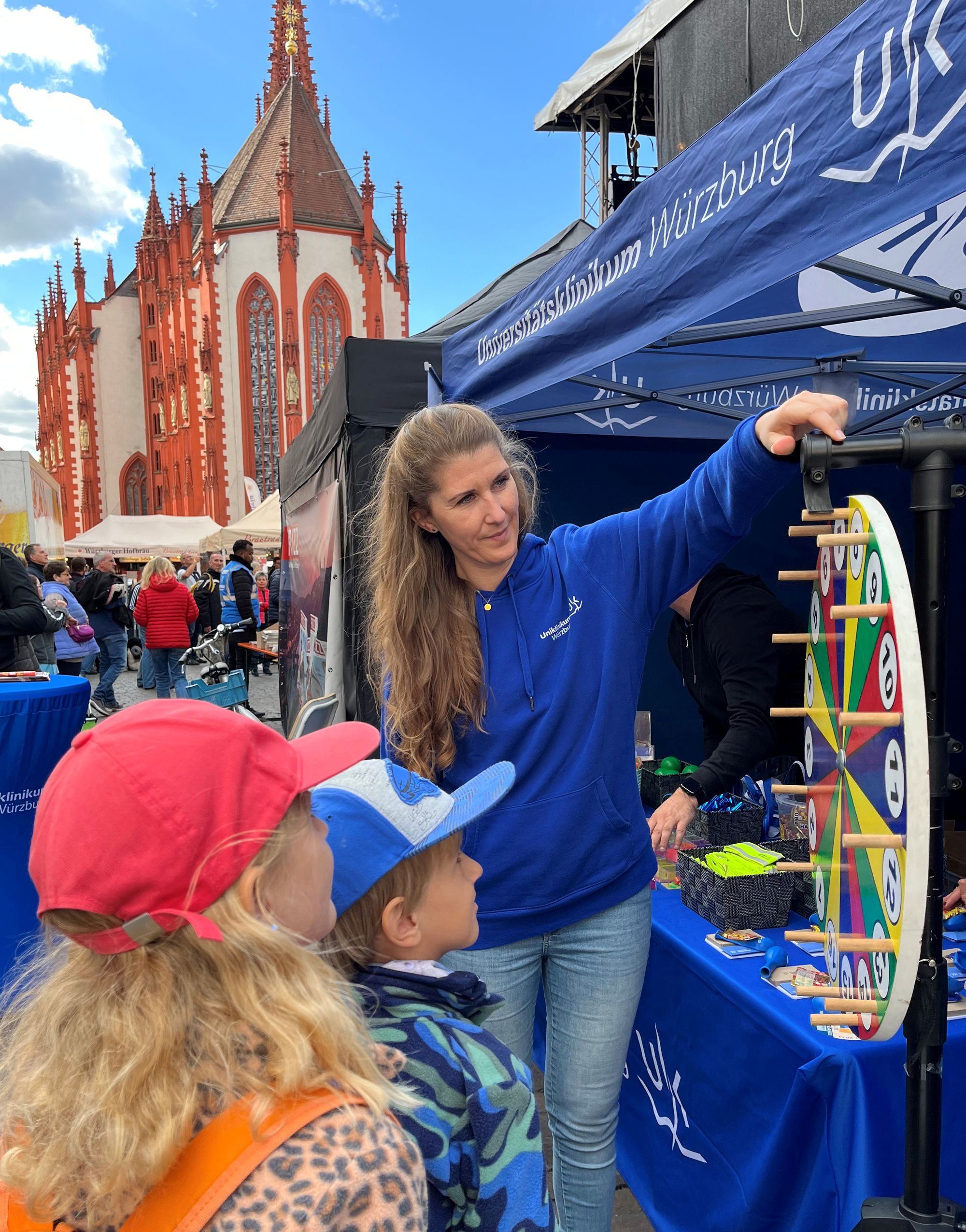 Am UKW-Stand steht eine Mitarbeiterin mit zwei Kindern am Glücksrad. Im Hintergrund ist die Kirche zu sehen.