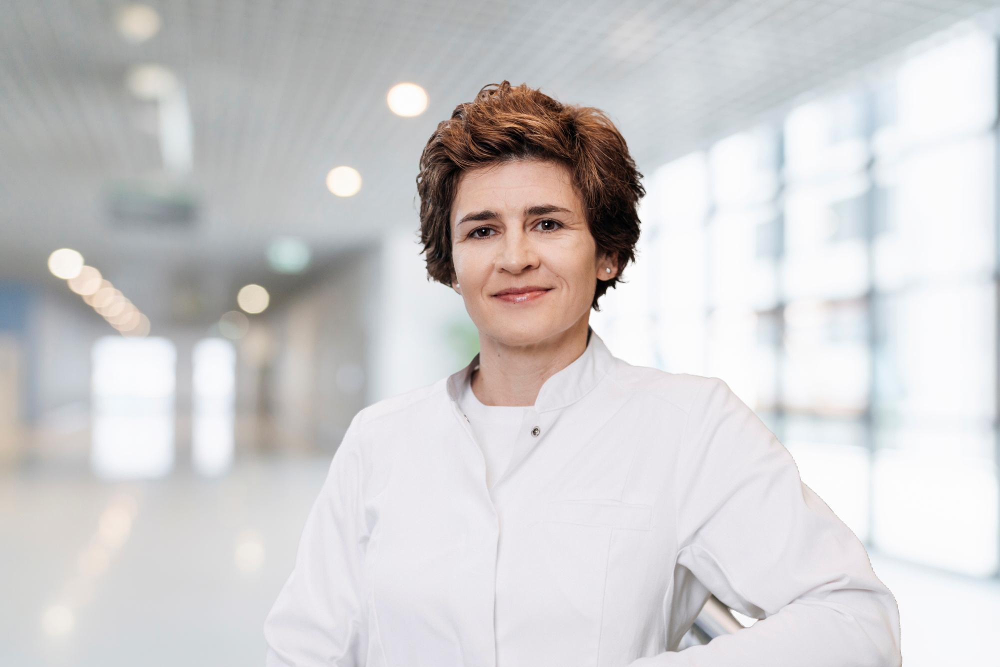 Anke Bergmann steht in weißem Kittel in einem Flur, der Hintergrund ist sehr unscharf. Das Bild ist im Querformat, Frau Bergmann ist bis zum Bauchnabel zu sehen. 