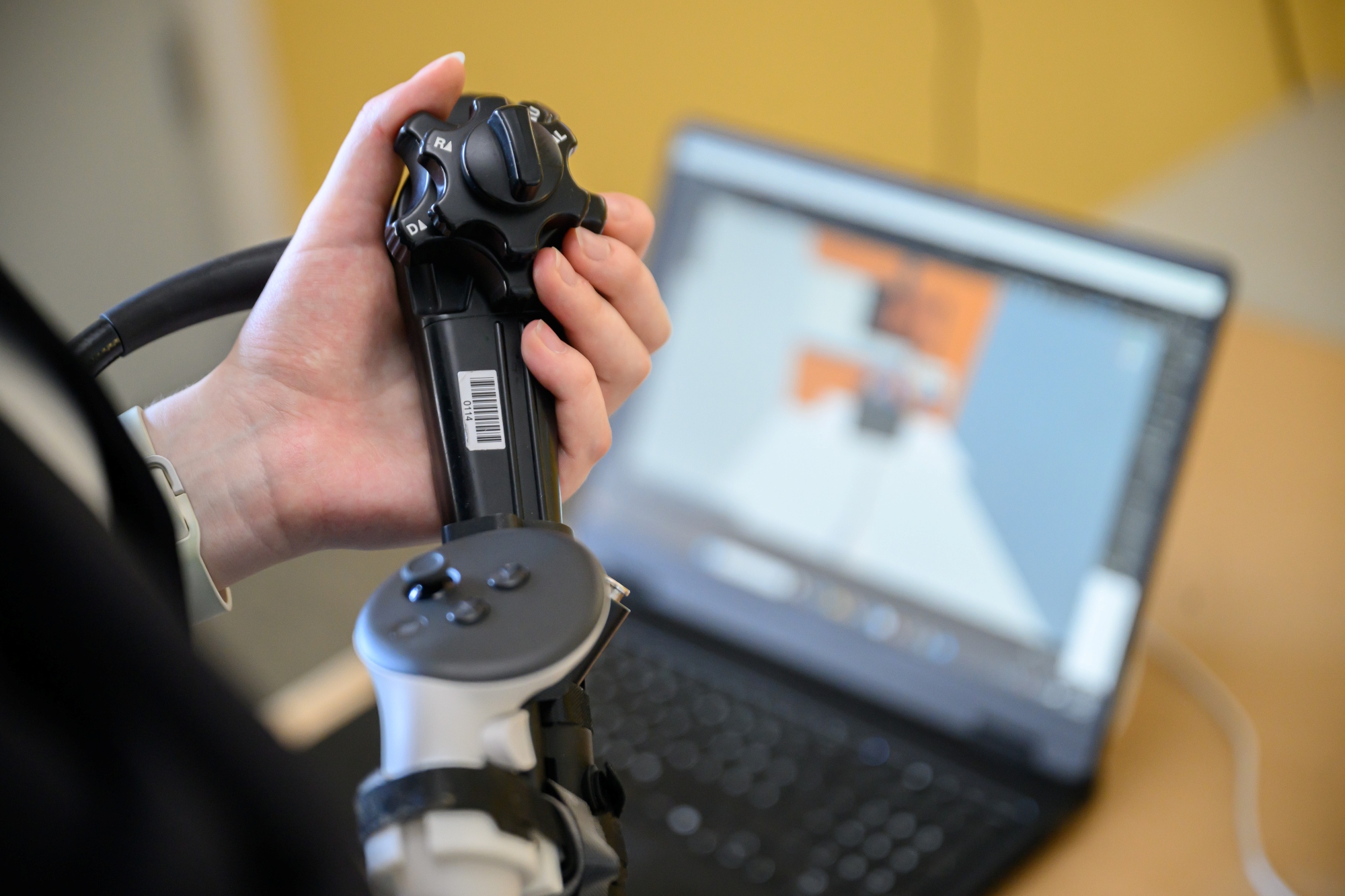 Nahaufnahme Endoskop, das in der Hand der Anwenderin liegt. Im Hintergrund der Bildschirm eines Laptops mit der virtuellen Welt. 