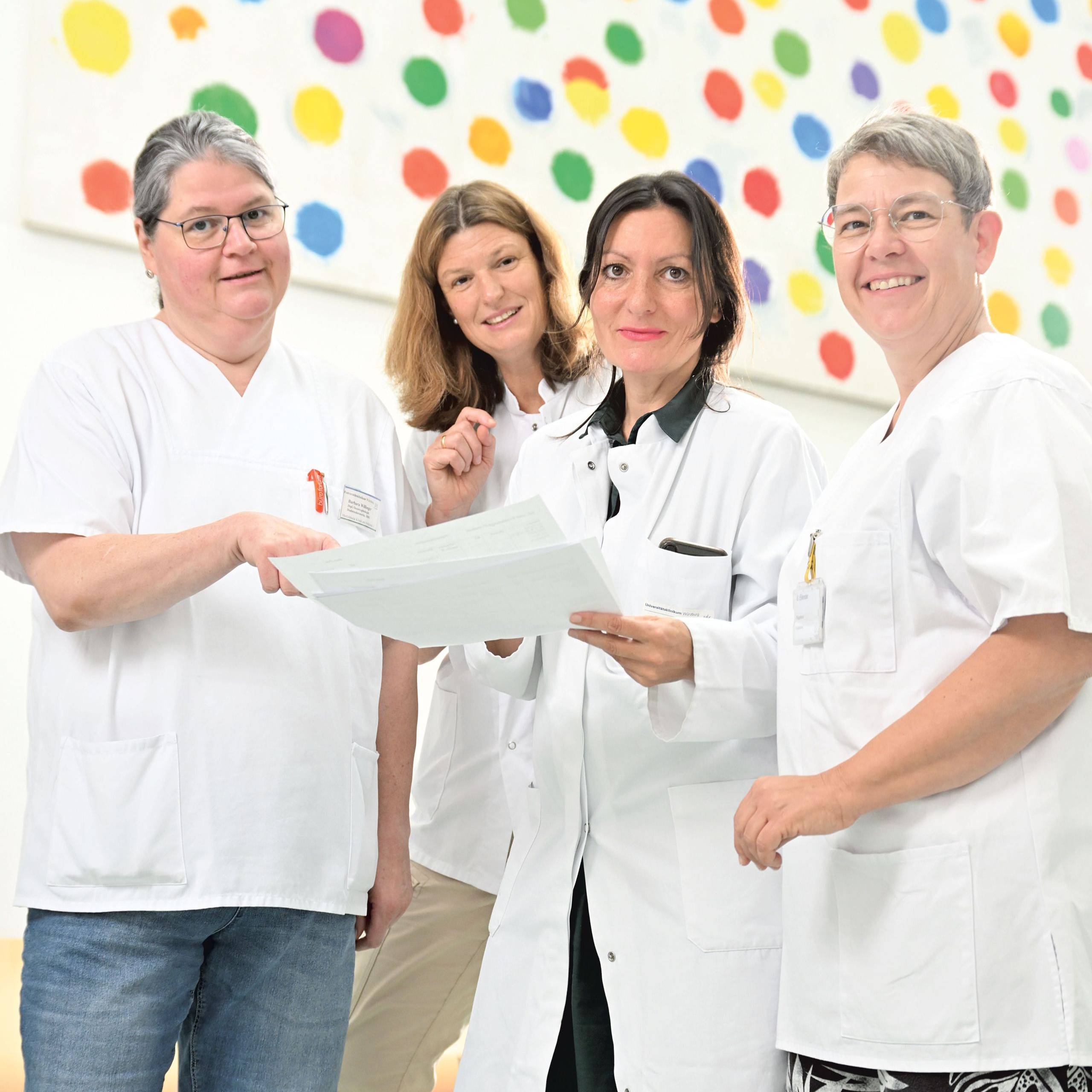 Vier Frauen, darunter Dr. Koschker, in weißen (Arzt- und Pflege-)Kleidung stehen auf einem Flur des UKW für eine Besprechung nebeneinander. 