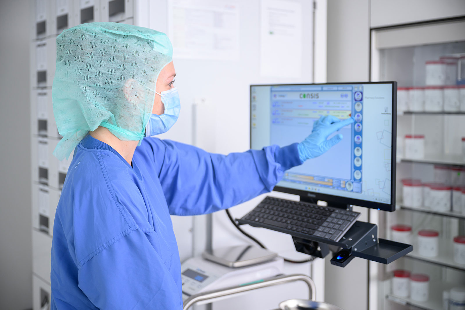 Mitarbeiterin in Reinraumkleidung (Haube, Maske, Handschuhe) bedient in Klinikapotheke unter sterilen Bedingungen einen Touchscreen.
