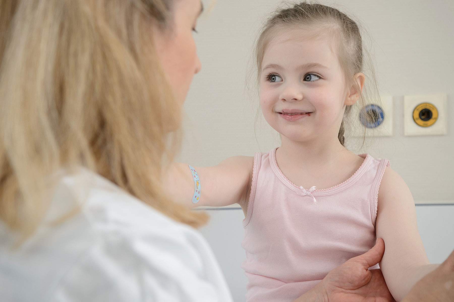Lächelndes Mädchen mit Pflaster am Arm im Gespräch mit einer Ärztin in der Kinderklinik.