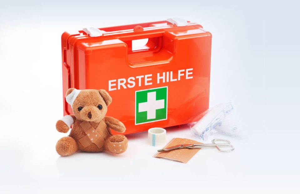 Ein orangefarbener Erste-Hilfe-Koffer. Im Vordergrund sind Verbandsmaterial und ein Teddybär mit Pflastern und Verbänden zu sehen.