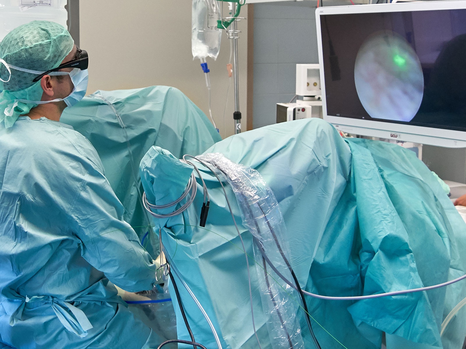 Ein Arzt sitzt in grüner OP-Kleidung vor einem abgedeckten Patienten, um eine endoskopische OP über die Harnröhre durchzuführen.