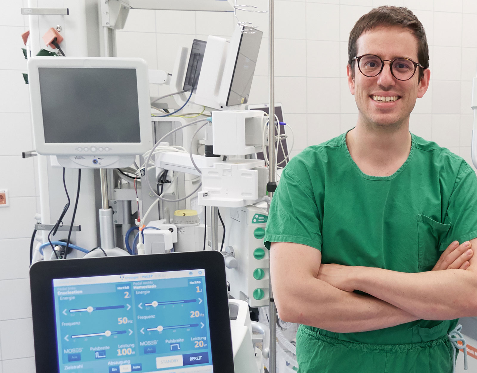 Dr. Charis Kalogirou, Oberarzt an der Klinik für Urologie und Kinderurologie steht in einem klinischen Behandlungsraum mit verschränkten Armen vor medizinischen Geräten und Monitoren und lächelt in die Kamera.