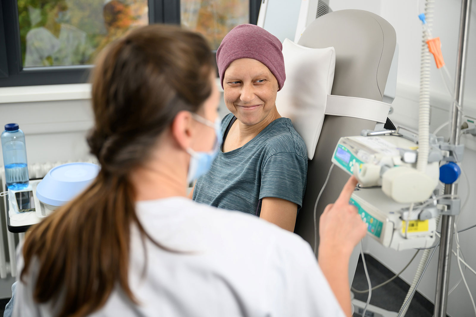 Patientin mit Kopftuch sitzt in einem Behandlungsstuhl und lächelt eine medizinische Fachkraft an, die an einem Infusionsgerät neben ihr Einstellungen überprüft.
