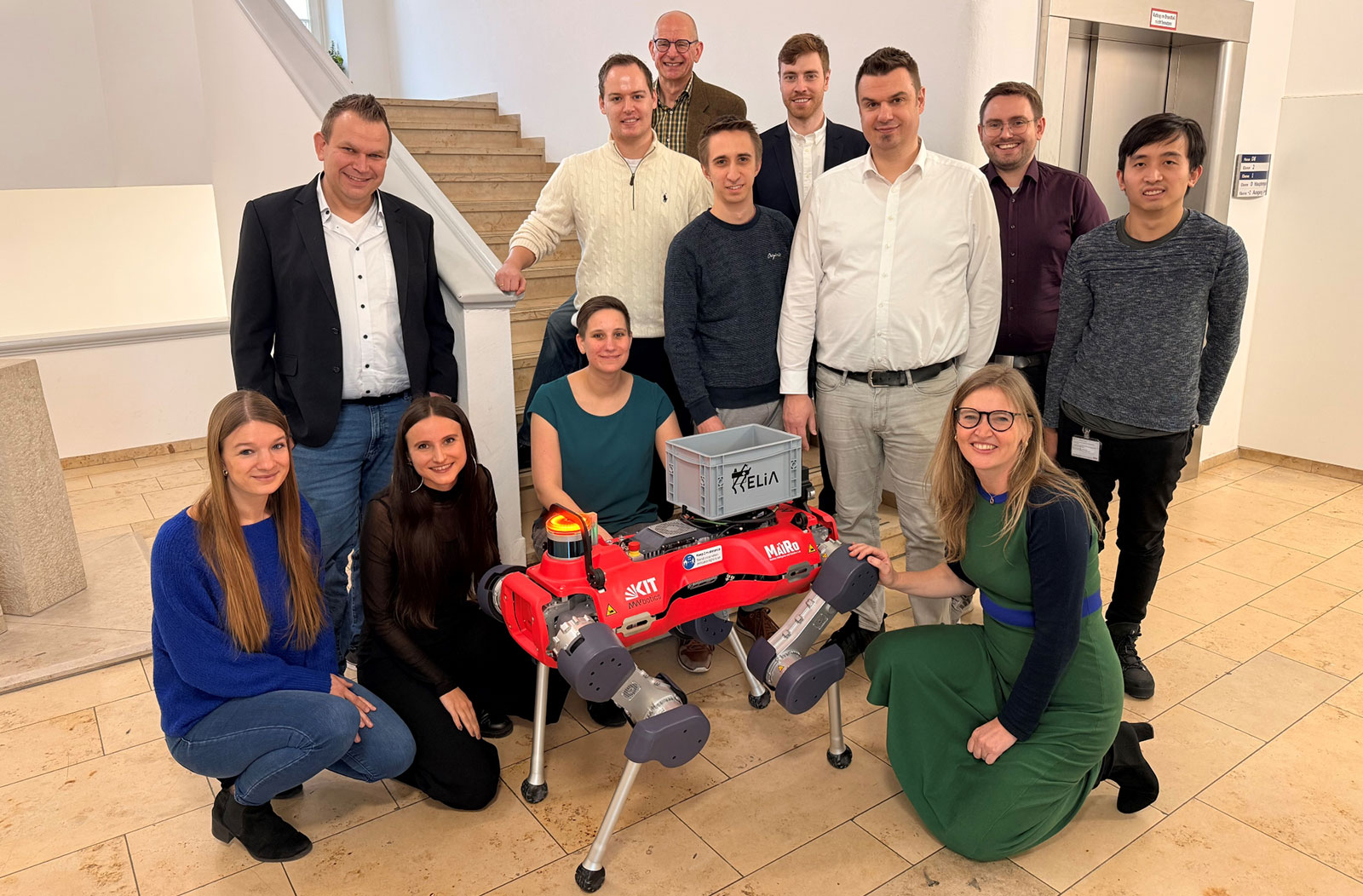 Eine Gruppe von Menschen steht um den Roboterhund HELIA. Im Vordergrund kniet Prof. Dr. Astrid Schmieder.