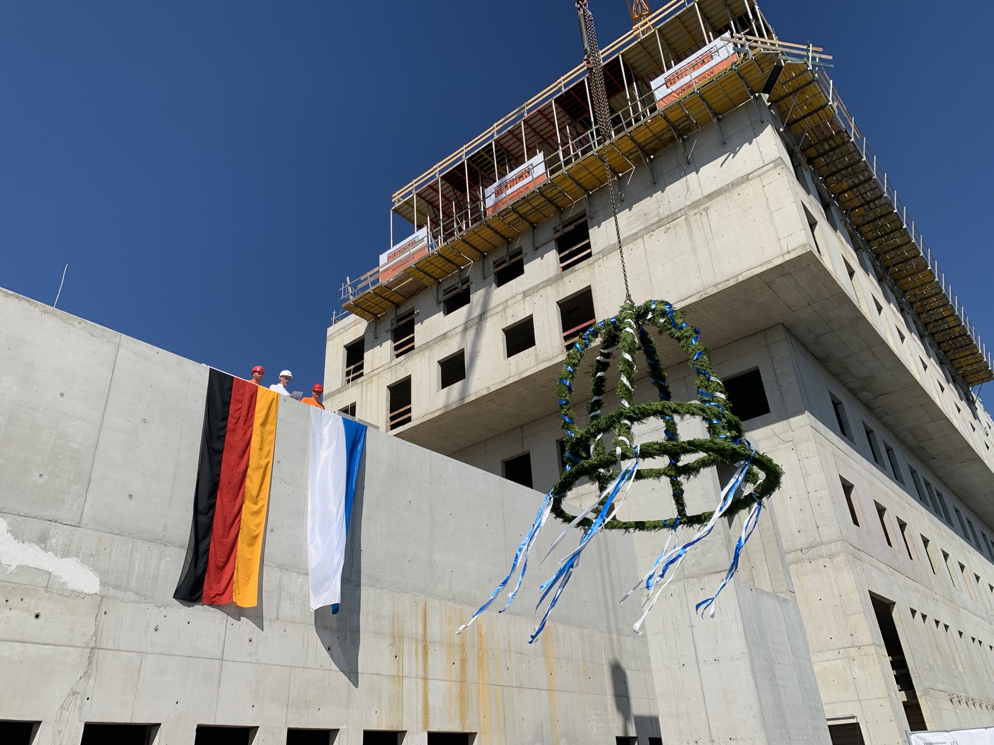 Ein Foto vom Richtfest des Neubaus der Strahlentherapie. Zu sehen ist ein Beton-Rohbau der sich auf der rechten Bildhälfte mehrere Geschosse erhebt. Auf der linken Bildhälfte sind die Deutschland-Flagge und die Bayrische Flagge zu sehen. Zentral im Bild ist ein Geschmückter Richtkranz mit blauen und weißen Bändern.