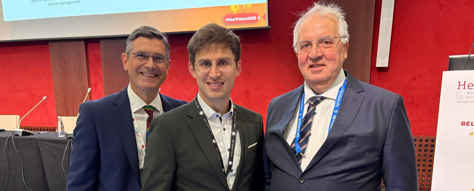 Die drei Wissenschaftler stehen in Anzügen vor der Bühne des Kongresses. 