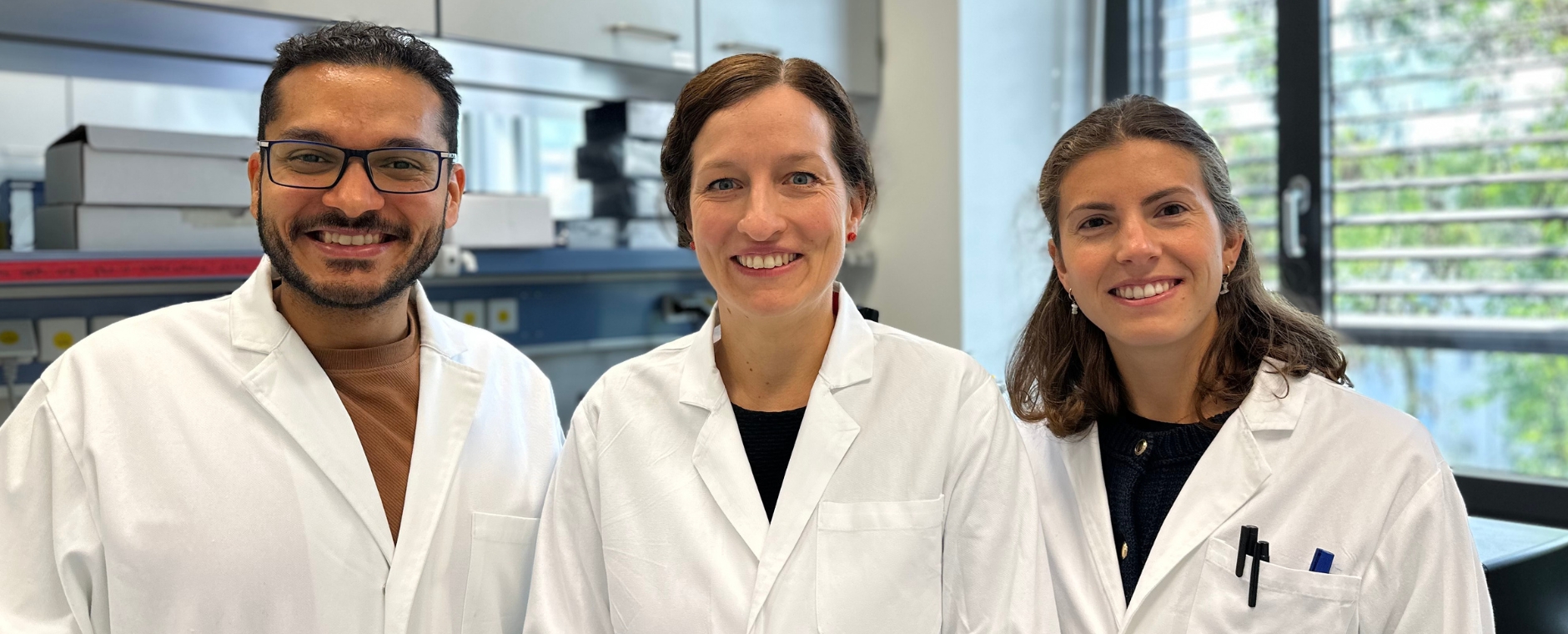 Helal, Riedel und Mattavelli Das Studienteam steht nebeneinander im Labor, alle tragen weiße Kittel und lachen in die Kamera.